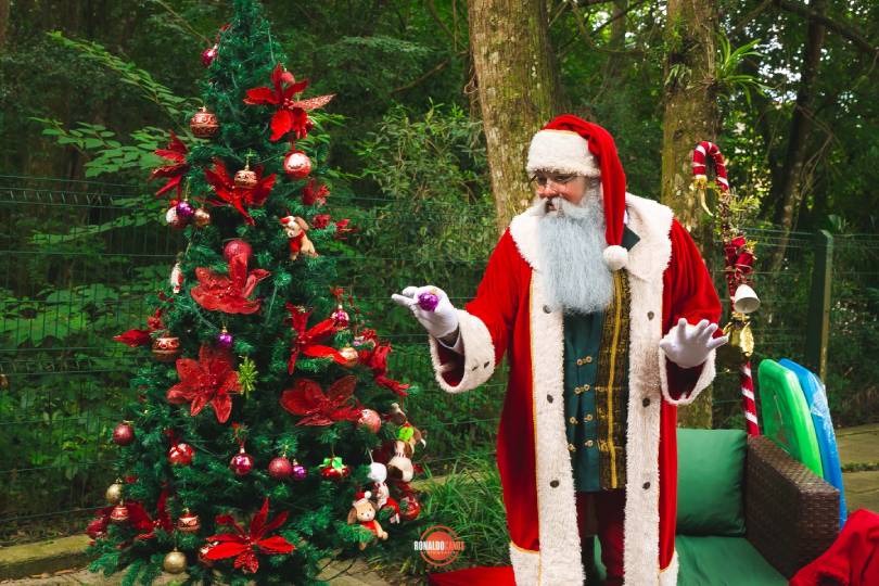 Papai Noel entregando um presente a duas crianças junto à árvore de Natal